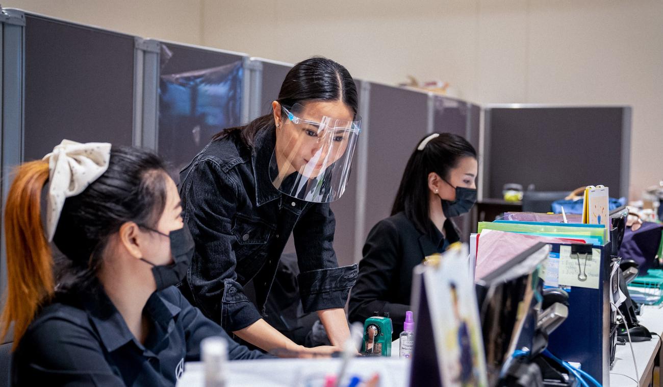 Trois jeunes femmes portant, pour l'une d'entre elle une visière de protection et, pour les deux autres, des masques de protection respiratoire, travaillent ensemble dans un bureau, devant des écrans d'ordinateurs.