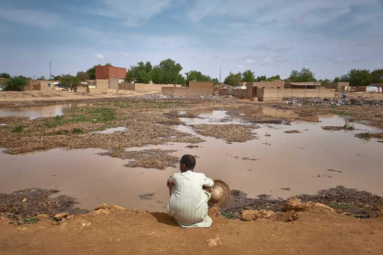 Un homme vêtu de blanc est assis par terre, dos à l'objectif, au bord d'un terrain inondé derrière lequel s'élèvent quelques constructions rudimentaires.