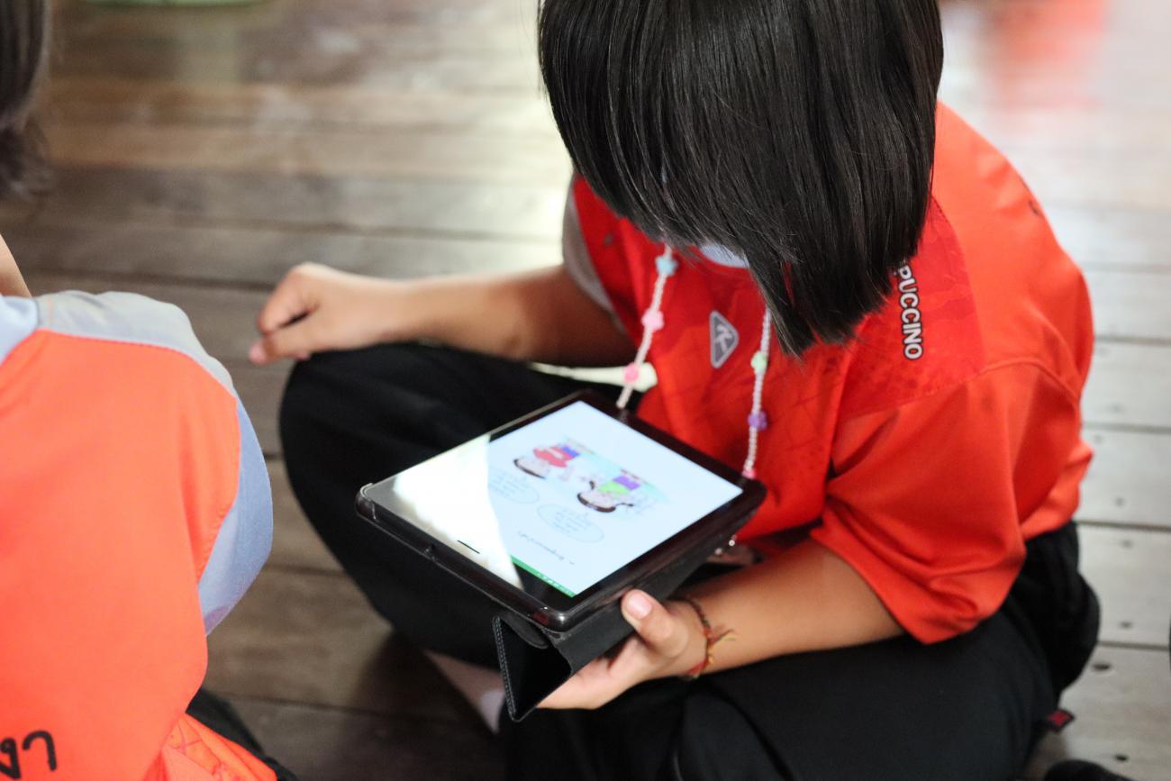 Un enfant vêtu d'un t-shirt orange foncé et portant une coupe de cheveux au carré est assis sur le sol en tailleur et regarde des dessins sur une tablette.