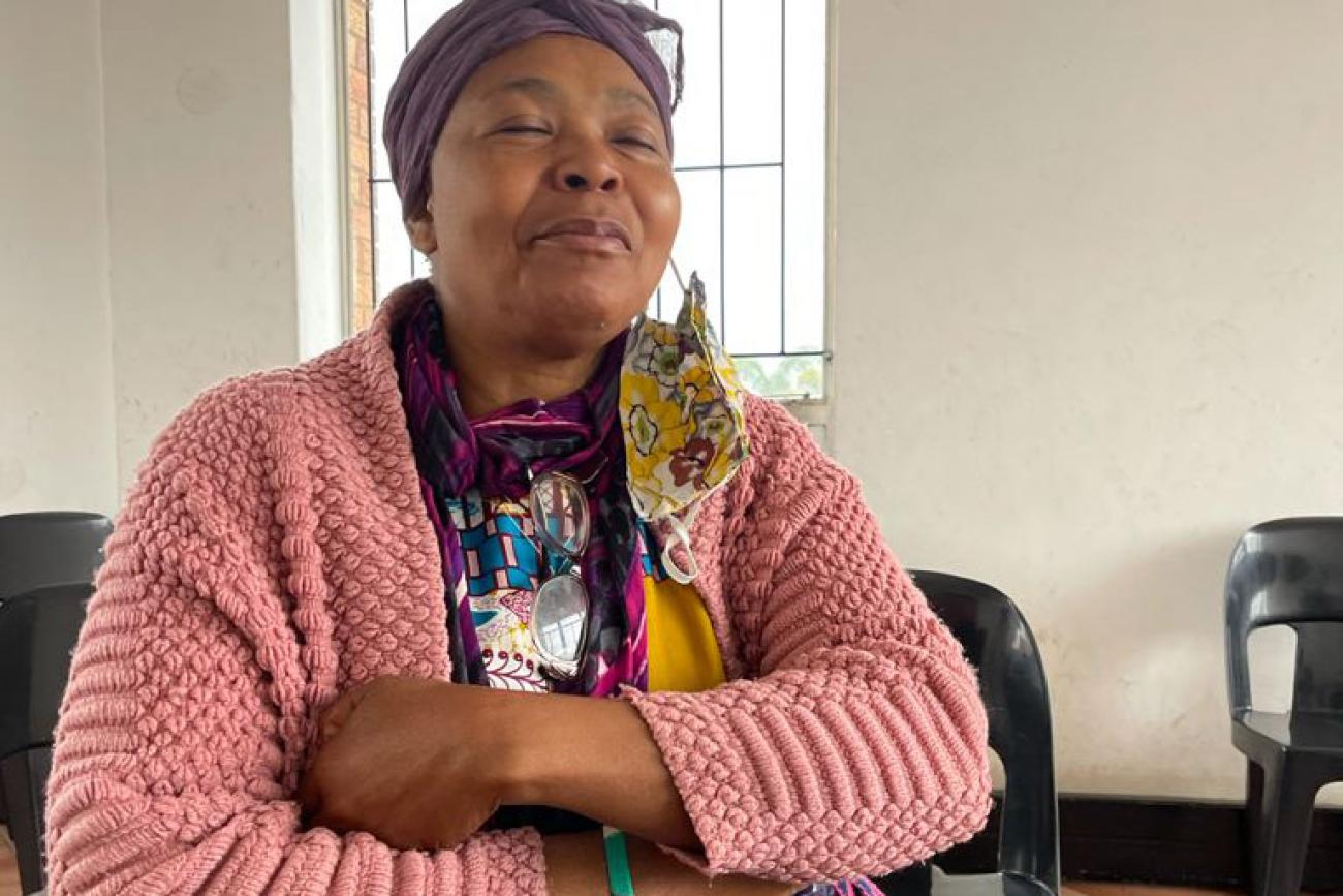 امرأة كبيرة في السن ترتدي سترة وردية تجلس في غرفة بيضاء وذراعيها مطويتان.