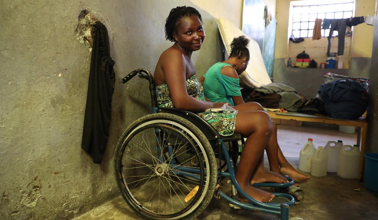 A woman sitting in a wheelchair smiles shyly at the camera, and another woman sits behind her looking away from the camera. 