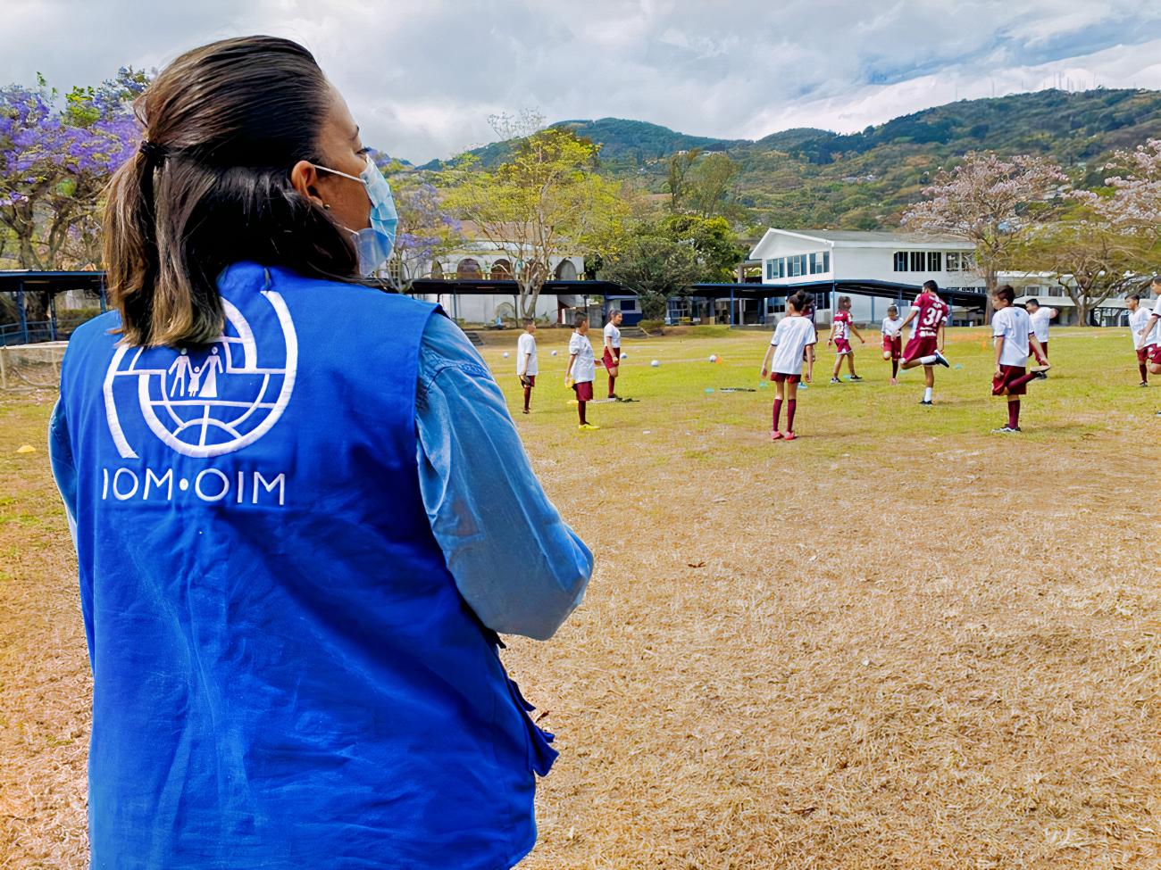 Au Costa Rica, une femme portant le gilet bleu de l’OIM et un masque chirurgical et faisant dos à la caméra regarde des enfants jouer au football sur un terrain en arrière-plan duquel on peut voir des collines verdoyantes.