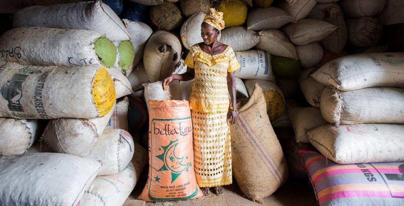 Une femme vêtue d’une robe traditionnelle africaine se tient debout dans un entrepôt à côté de nombreux sacs de semences.