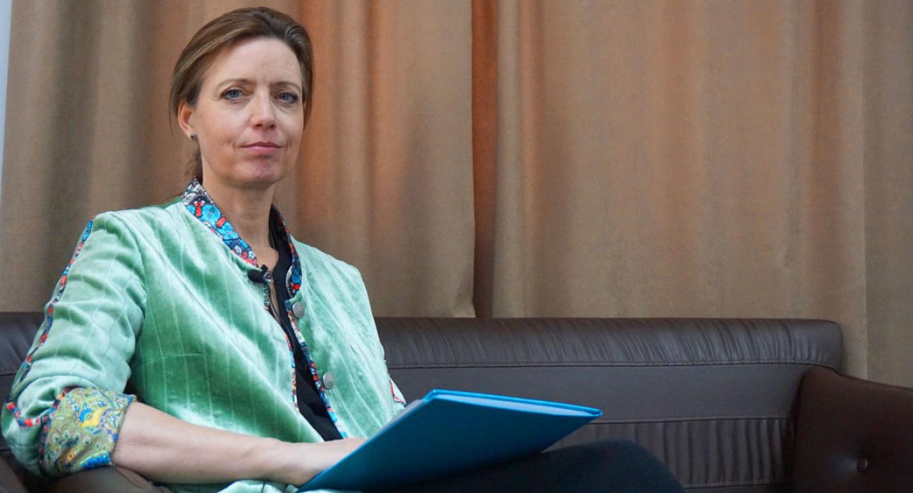 Женщина в бирюзовом пиджаке сидит на кресле с папкой в руках.