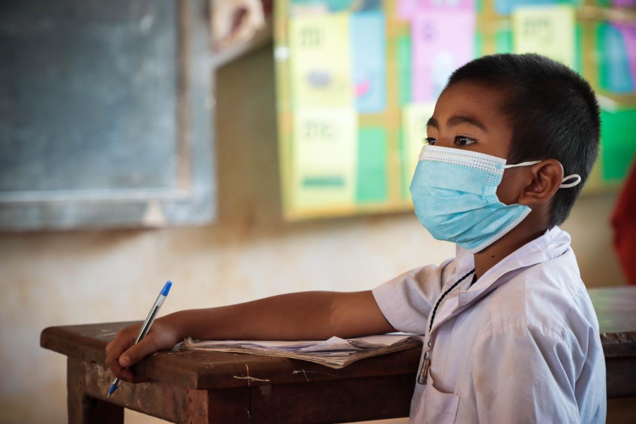 يجلس طالب في أحد الفصول الدراسية مرتديًا الكمامة ويمسك بقلم.