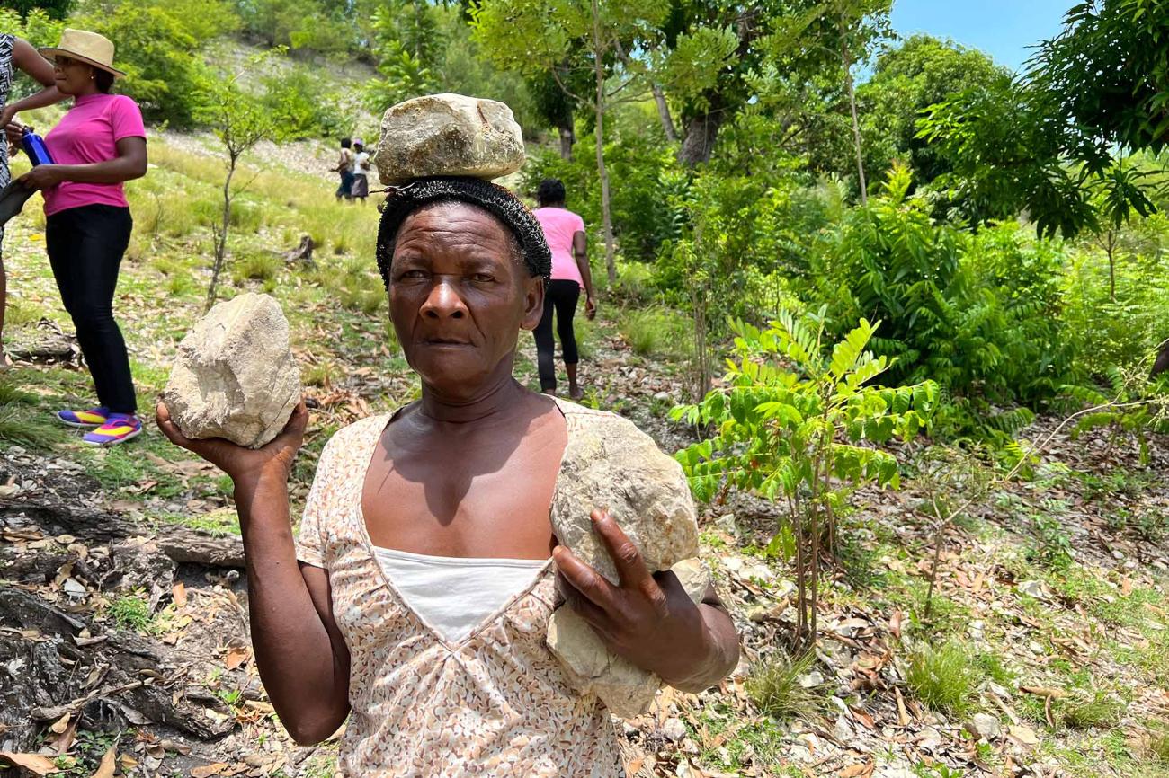 Portrait of a woman in a beige and white blouse, looking determinedly at the camera and she is part of a line of women carrying rocks in their hands and on their heads as they descend a ravine on a hillside in the southern Haiti.
