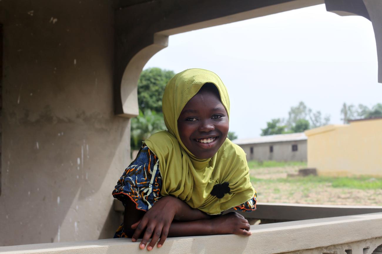 Une jeune fille portant un voile de couleur claire autour de la tête est penchée au-dessus d'un balcon dans une campagne togolaise et regarde en direction de la caméra en souriant.
