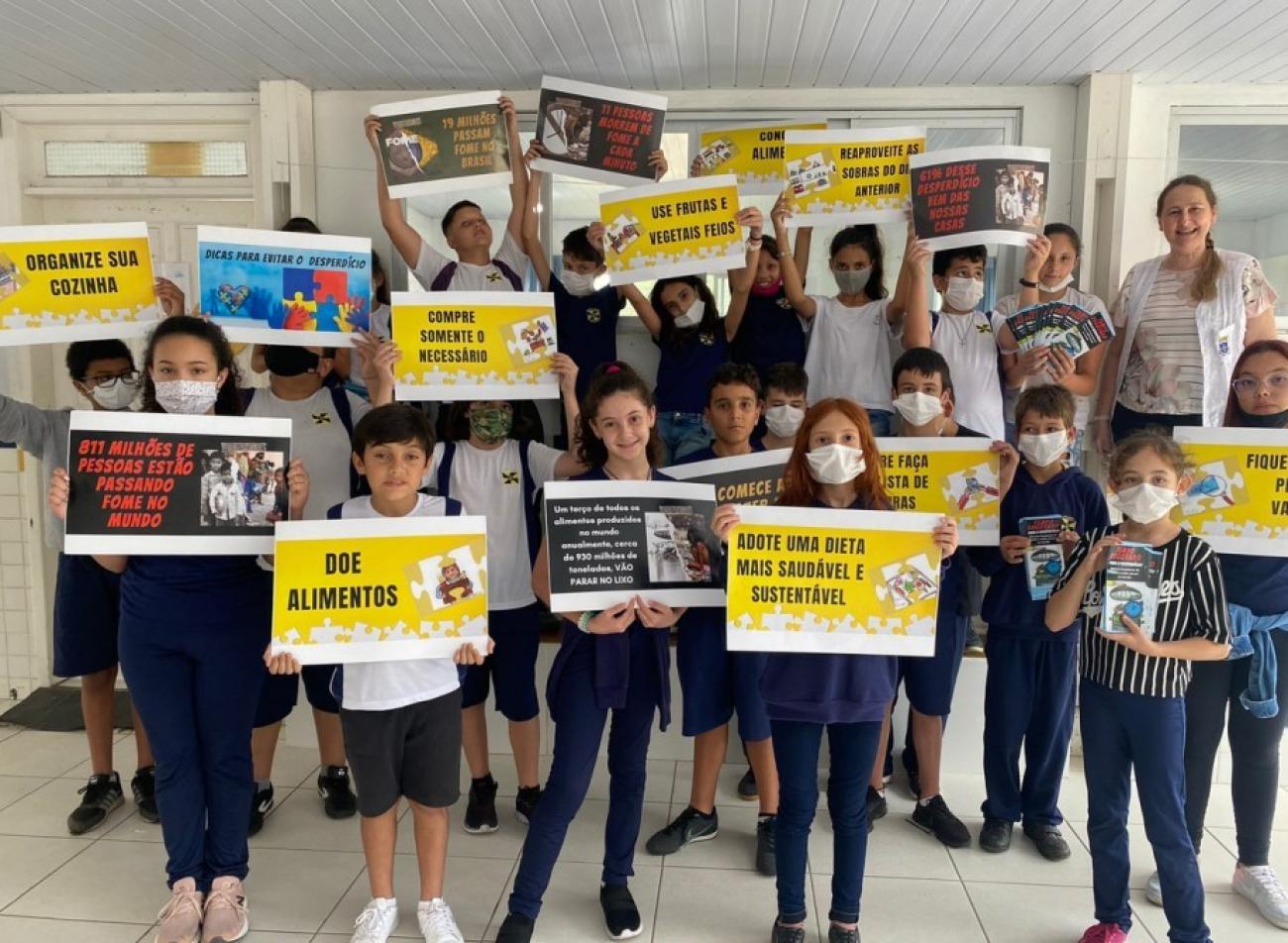 مجموعة من الطلاب في سن المدرسة الابتدائية مع معلمتهم. يقفون جنبًا إلى جنب ويعرضون بفخر أعمالهم المدرسية في مجال إدارة النفايات الغذائية.