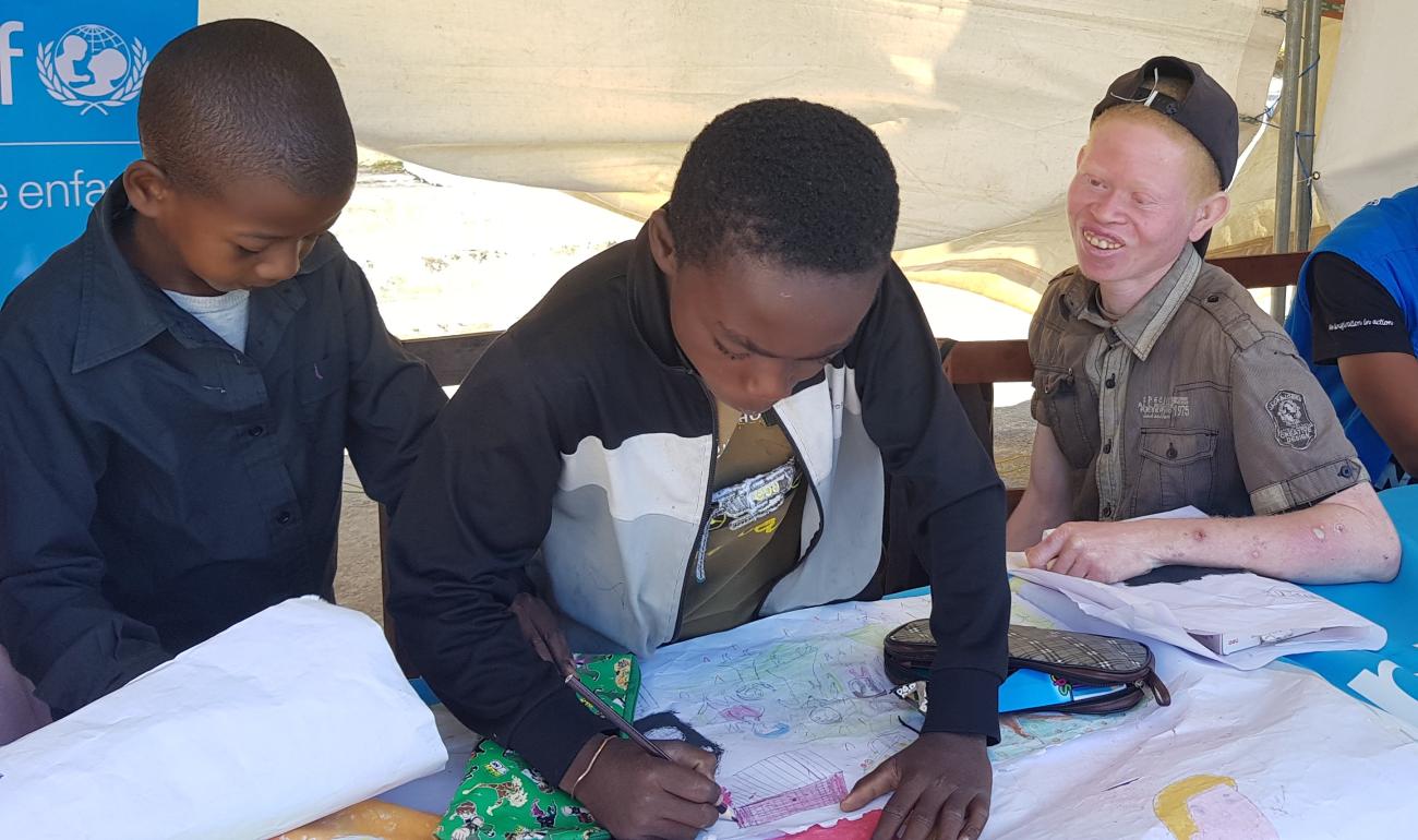 يدرس صبيان صغيران على طاولة تحت خيمة كبيرة أقامتها اليونيسف، بينما طفل ثالث يشاهد بابتسامة كبيرة.