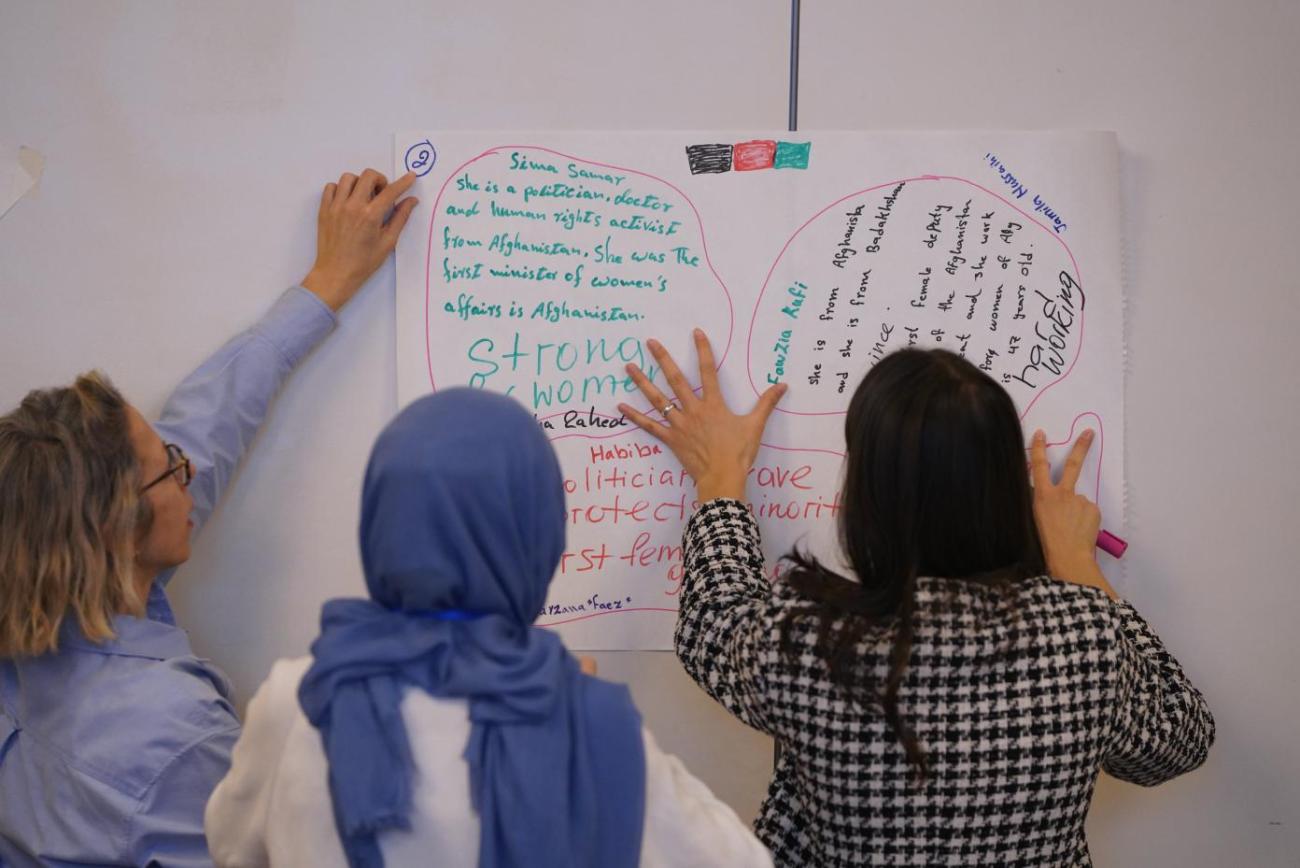 Three women work on a whiteboard and point to scribbled notes, with their backs facing the camera.