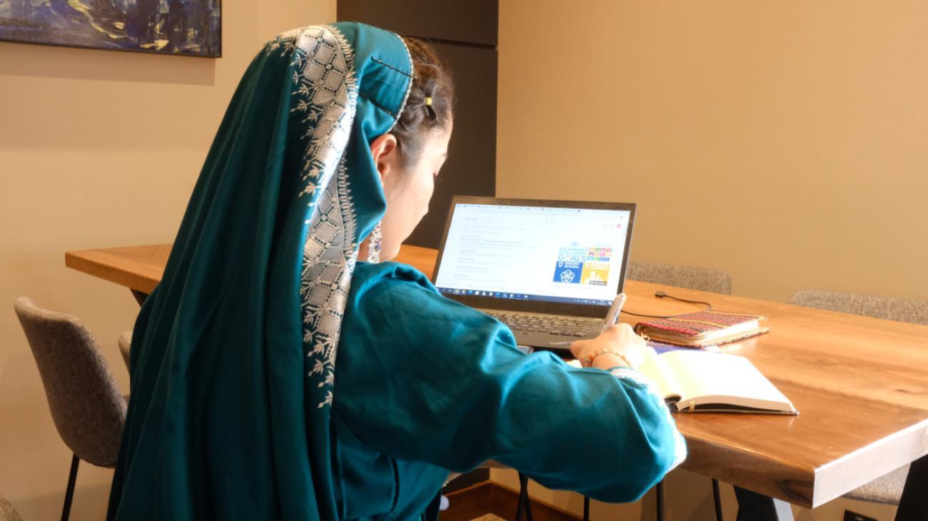 A female in a green satin-like dress with matching headscarf faces away from the camera and works on a laptop.