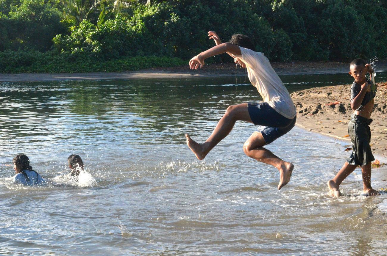 A group of young kids jump into a pool of water that appears like a lake with sand.