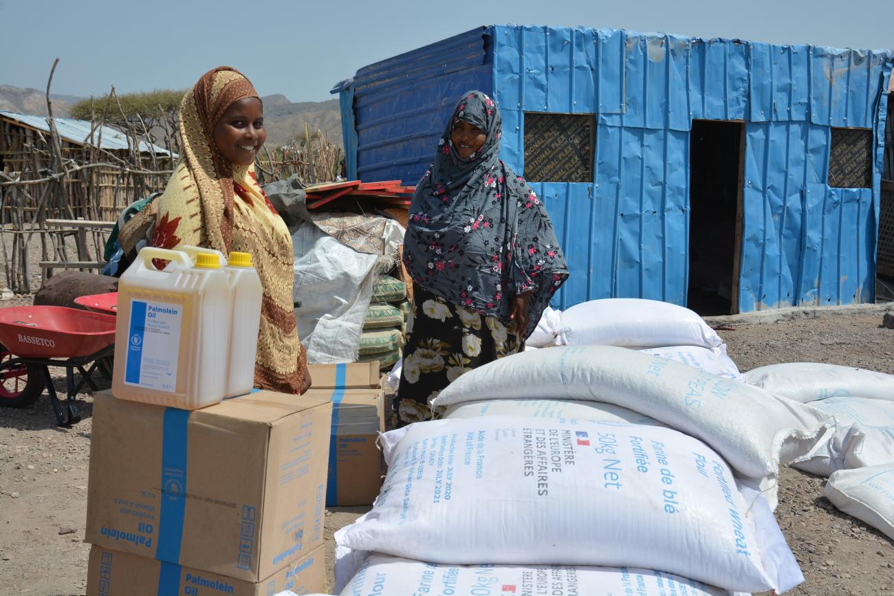 A woman in a brown dress and headscarf stands behind several boxes and cartons and sacks of emergency supplies. A blue cabin is in the background.