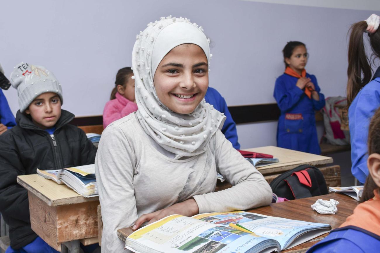 A girl in a white dress and headscarf sits at a school desk with a book open