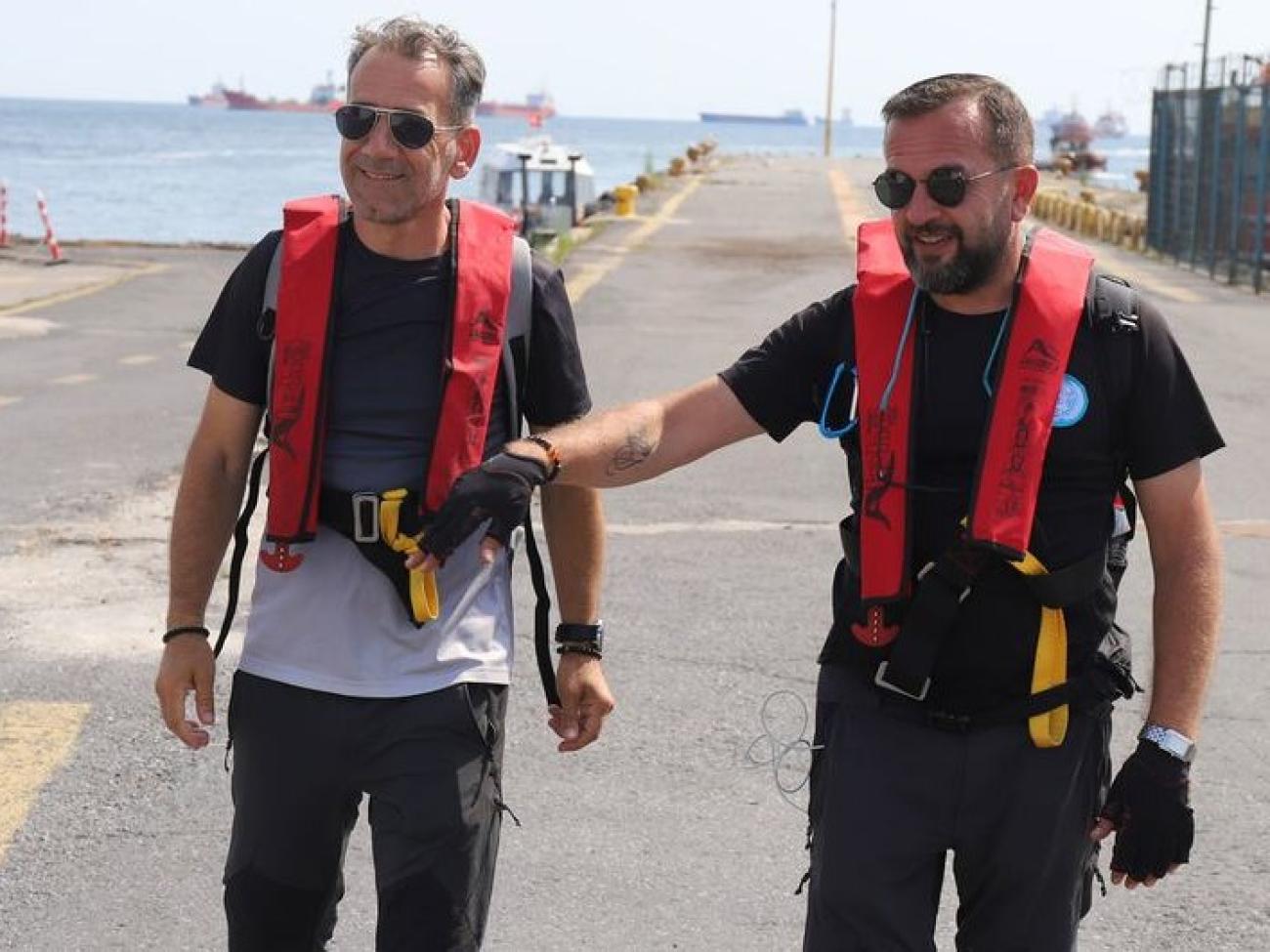 Two UN inspectors in red vests walk along an industrial pier.