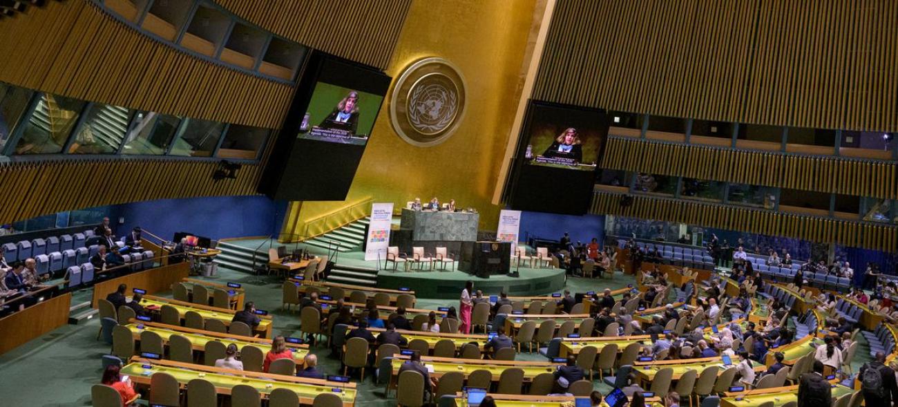 The General Assembly hall in the UN features rows and a podium with a gold backdrop.