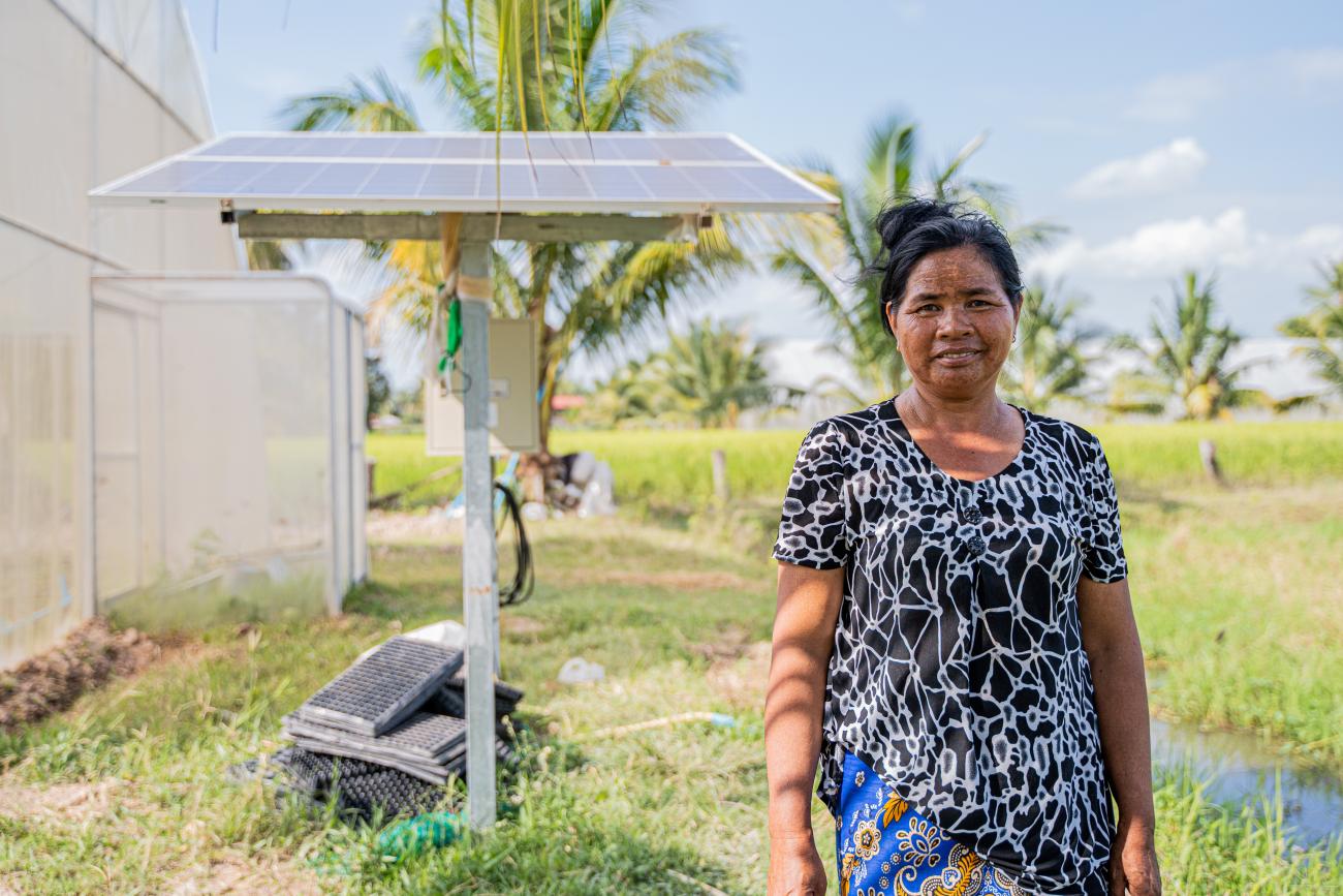 A woman in a black printed dress stands in front of a solar panel installed in a green field in rural Cambodia