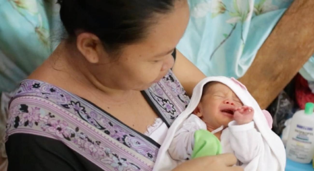 A woman in a purple dress cradles her new born baby in a white swaddle