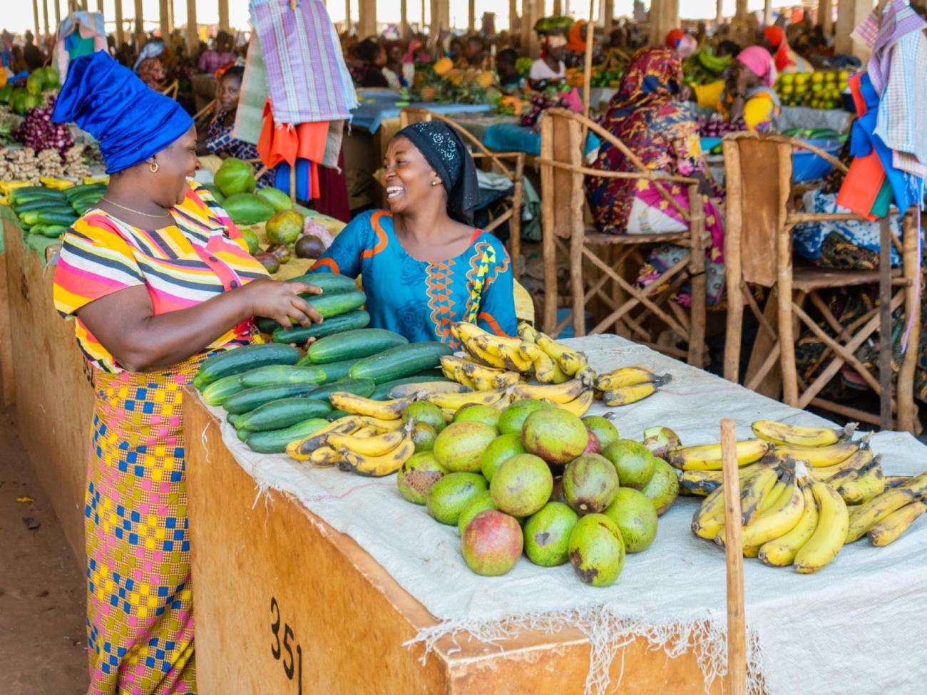Two women in colourful clothes laugh with each other in a market place, behind a stall selling mangoes and bananas