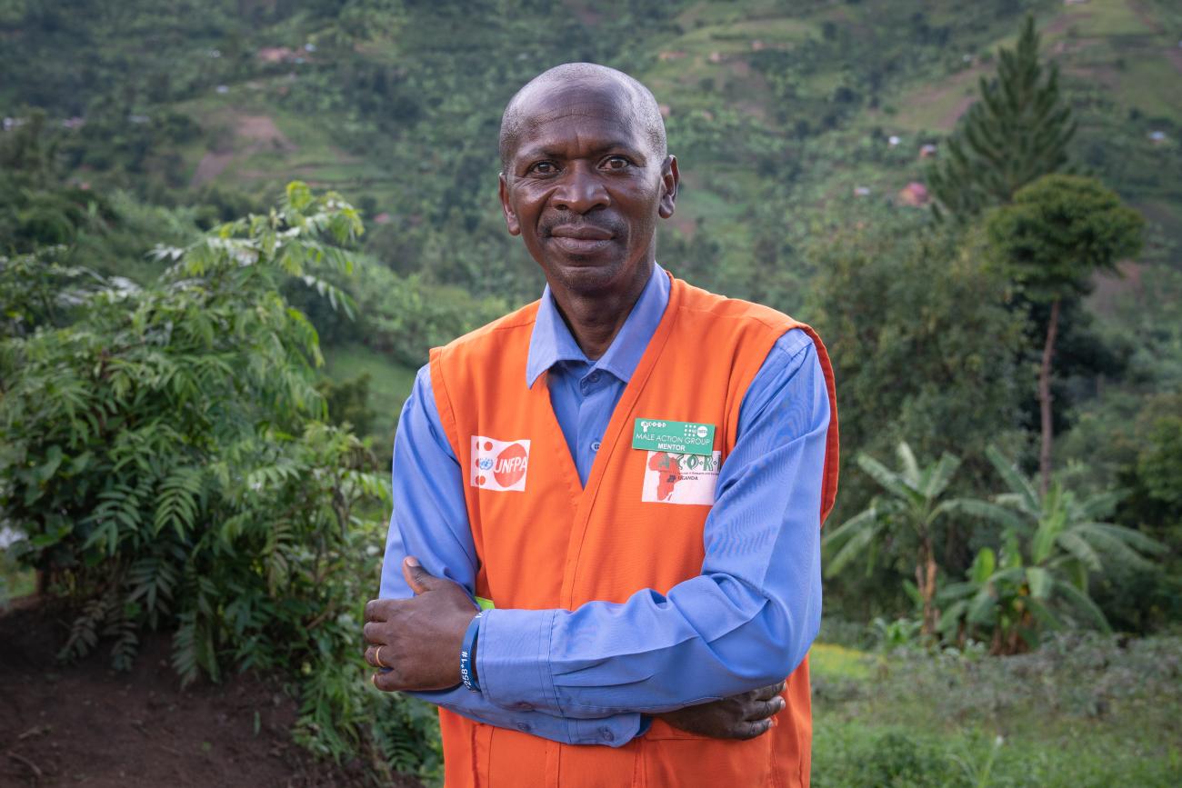 A man in a blue shirt and orange vest stands with his arms crossed in front of a green leafy background.