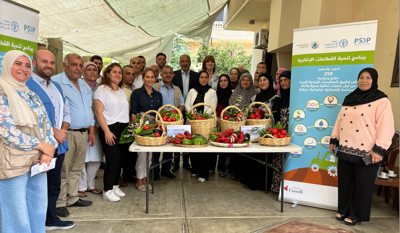 A group of people, men and women, in different coloured clothes pose for a picture behind a table where various fruits and vegetables are displayed.