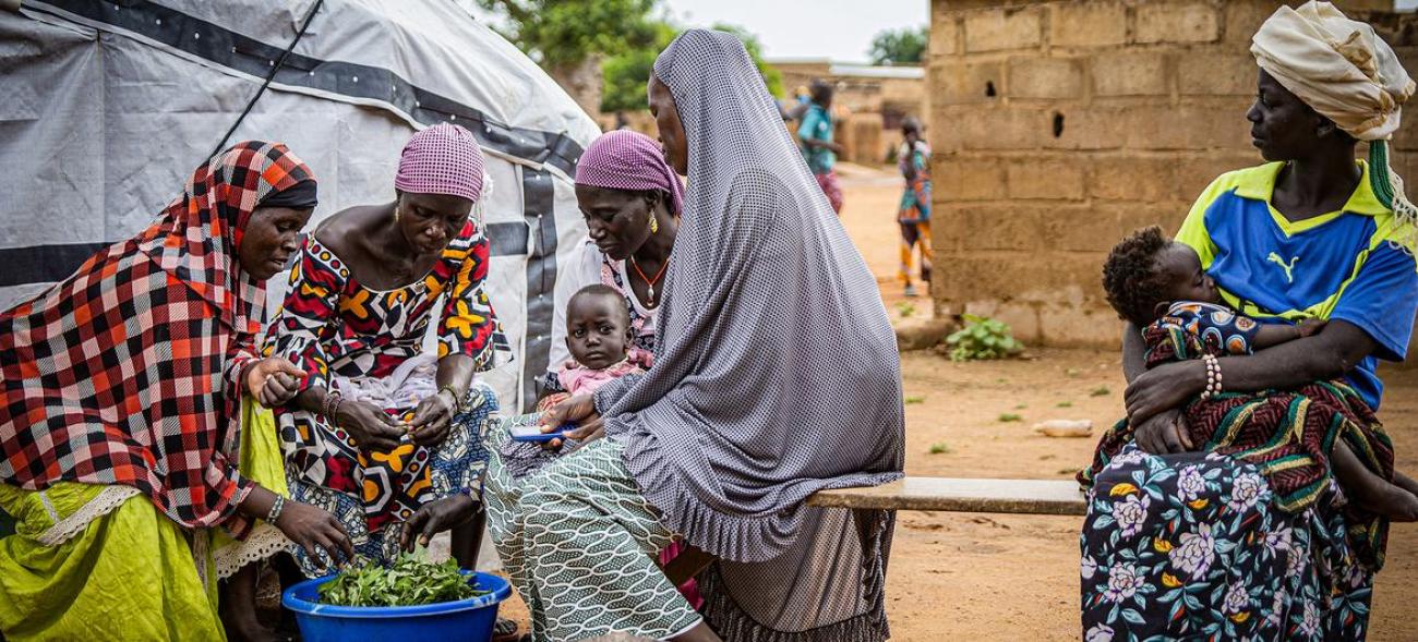 Women with head coverings sit on a bench with children on their laps, in a red-dusty setting.