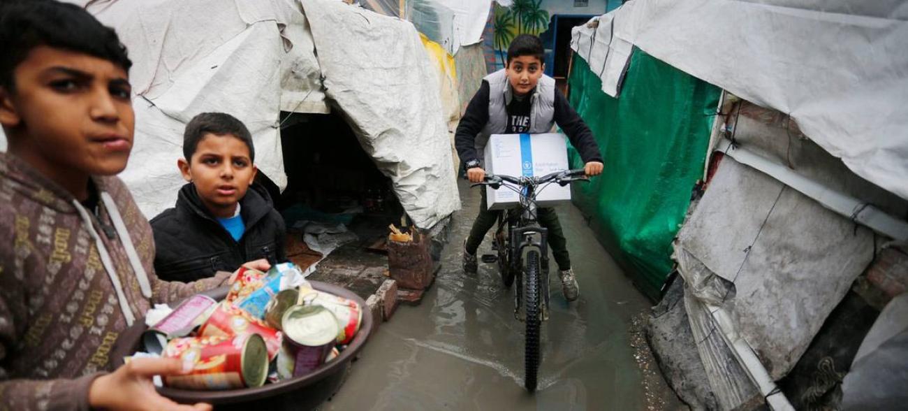 A picture of three children, one of them is on a bicycle, two of them hold baskets of food assistance