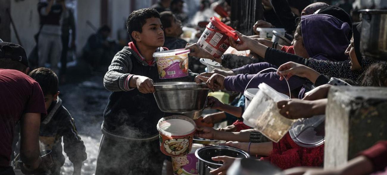 A young boy hands a large metal bowl, presumably with food, to people, including youth, lined up for food and water.