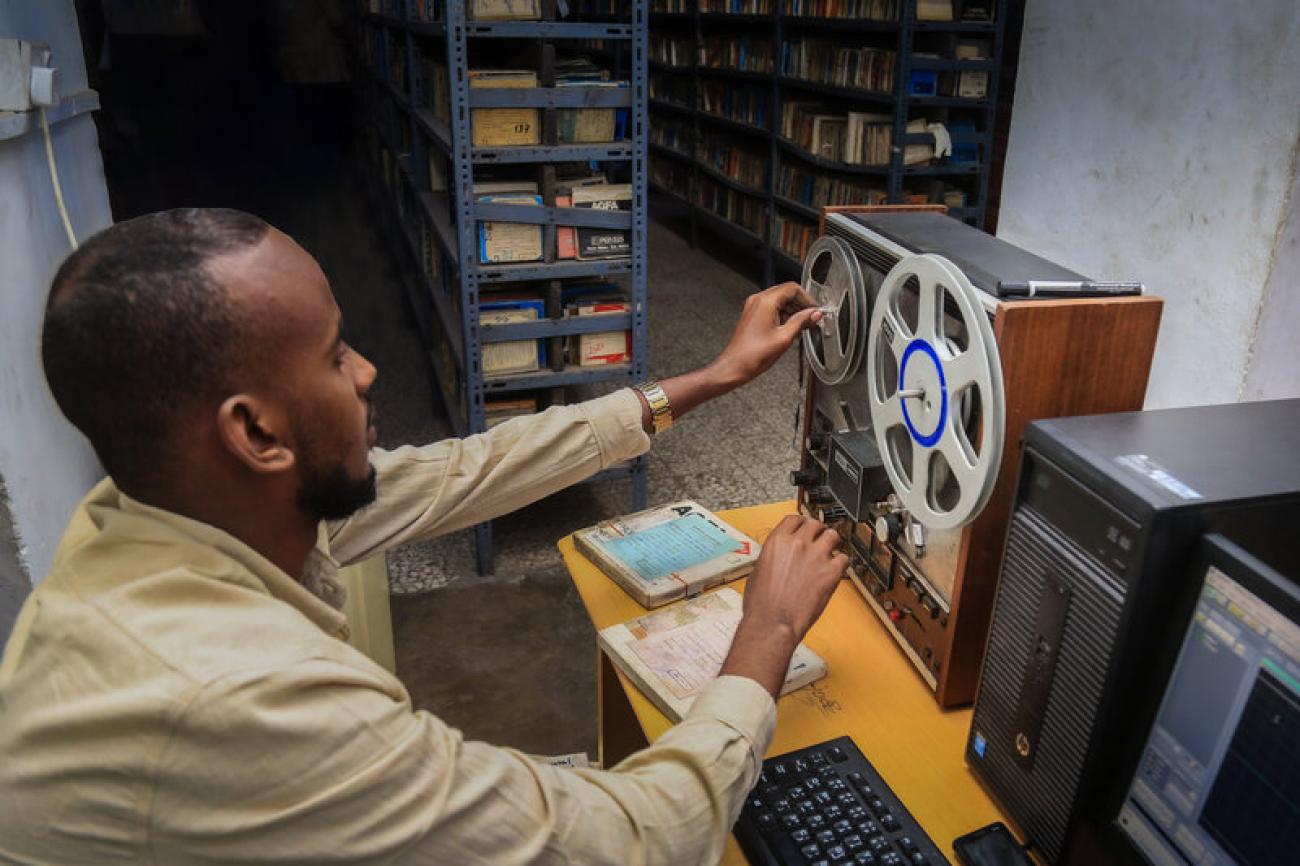 A man in a greenish brown shirt turns a record with his hands in a dark room