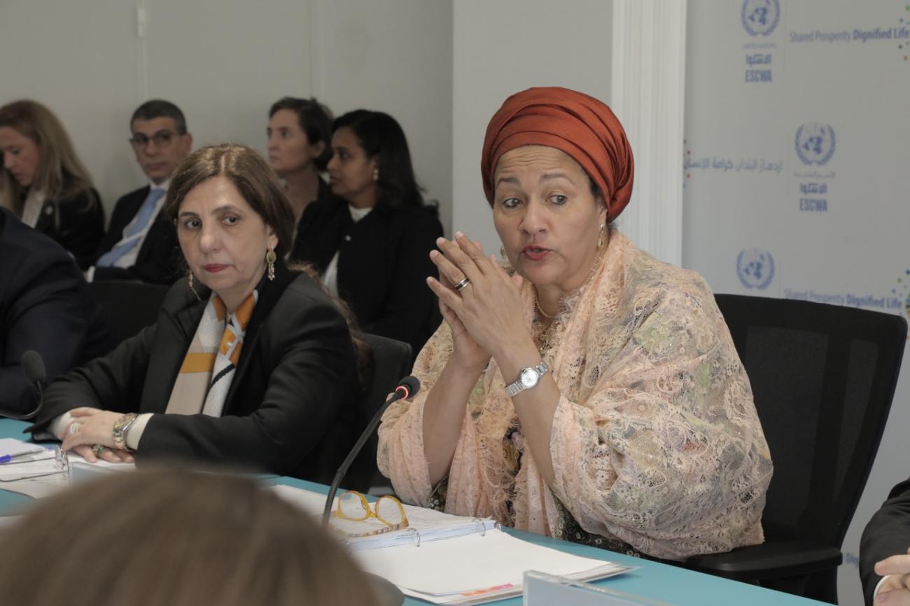 Two women, one in a red headdress and another in a black suit sit a table where they appear to be addressing other people.