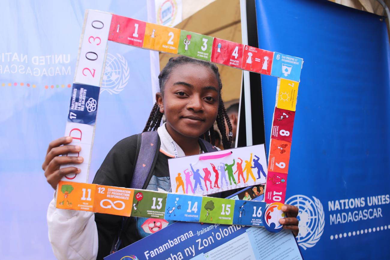 A young girl in a school uniform holds a frame with the SDGs, she looks at the camera and smiles