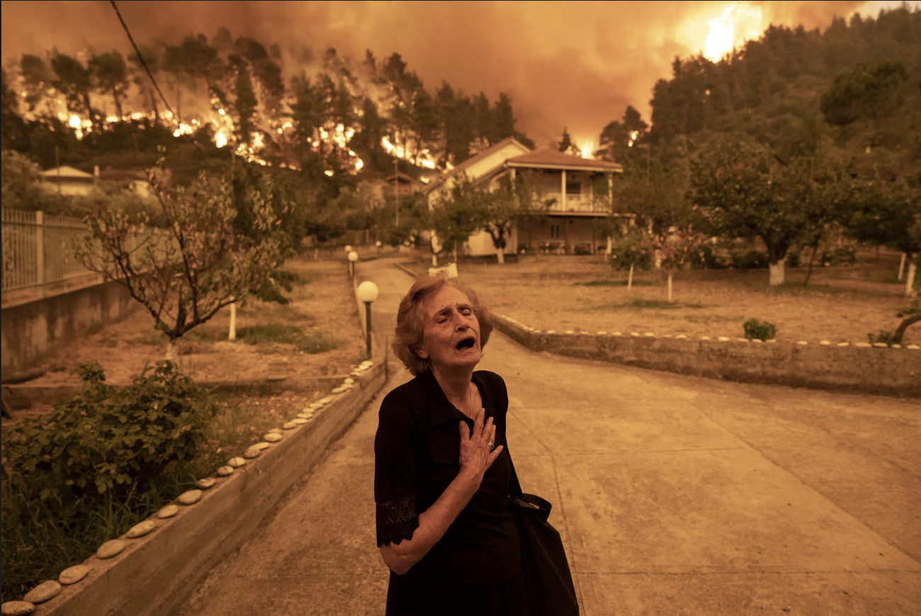 A woman in a black dress stands in the middle of a road holding her hand to her heart while there are forest fires in the background