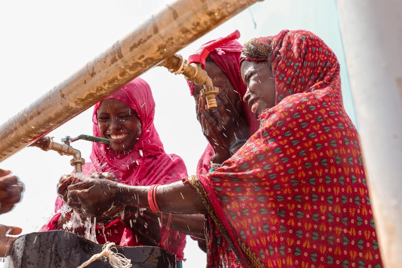 A young girl in a vibrant red garment with yellow and green patterns is collecting water from an outdoor tap into a black container, causing water to splash around. The tap is mounted on large, aged white pipes with visible rust, and the clear sky suggests a sunny day. Two other girls are partially visible. 