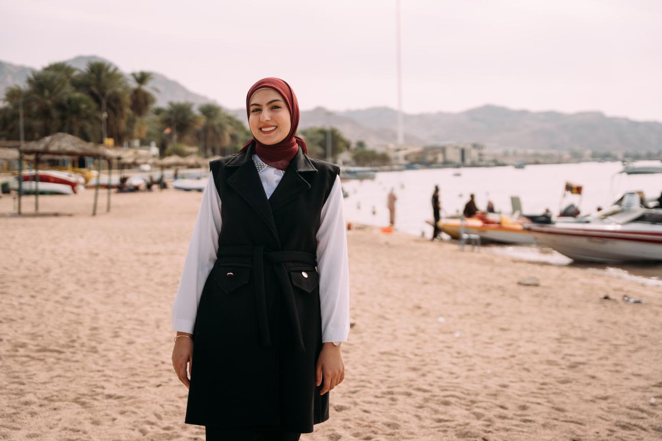 A woman in a black dress and red headcarf stands on a sandy beach.