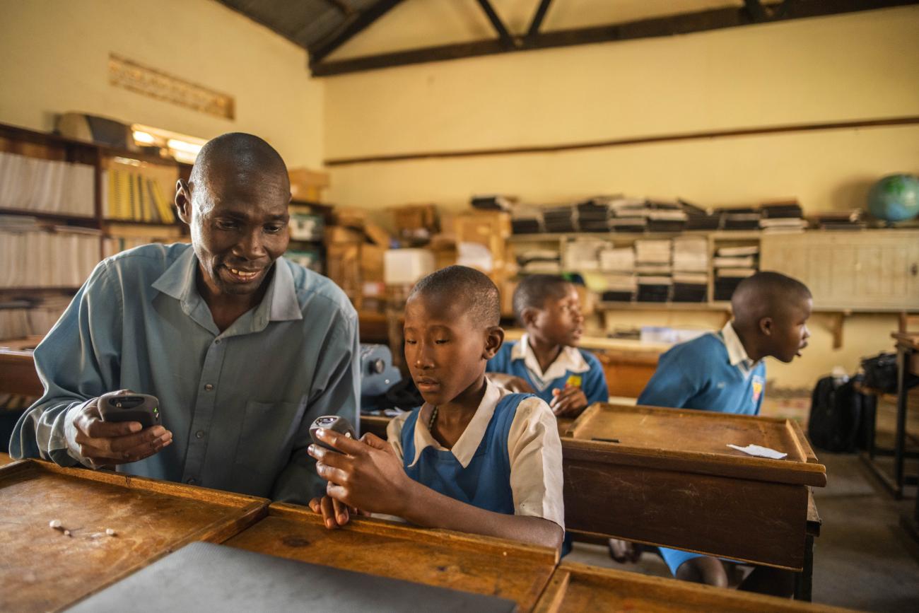A man in a grey shirt, a teacher, helps a young boy in a school uniform with a mobile device in a classroom.