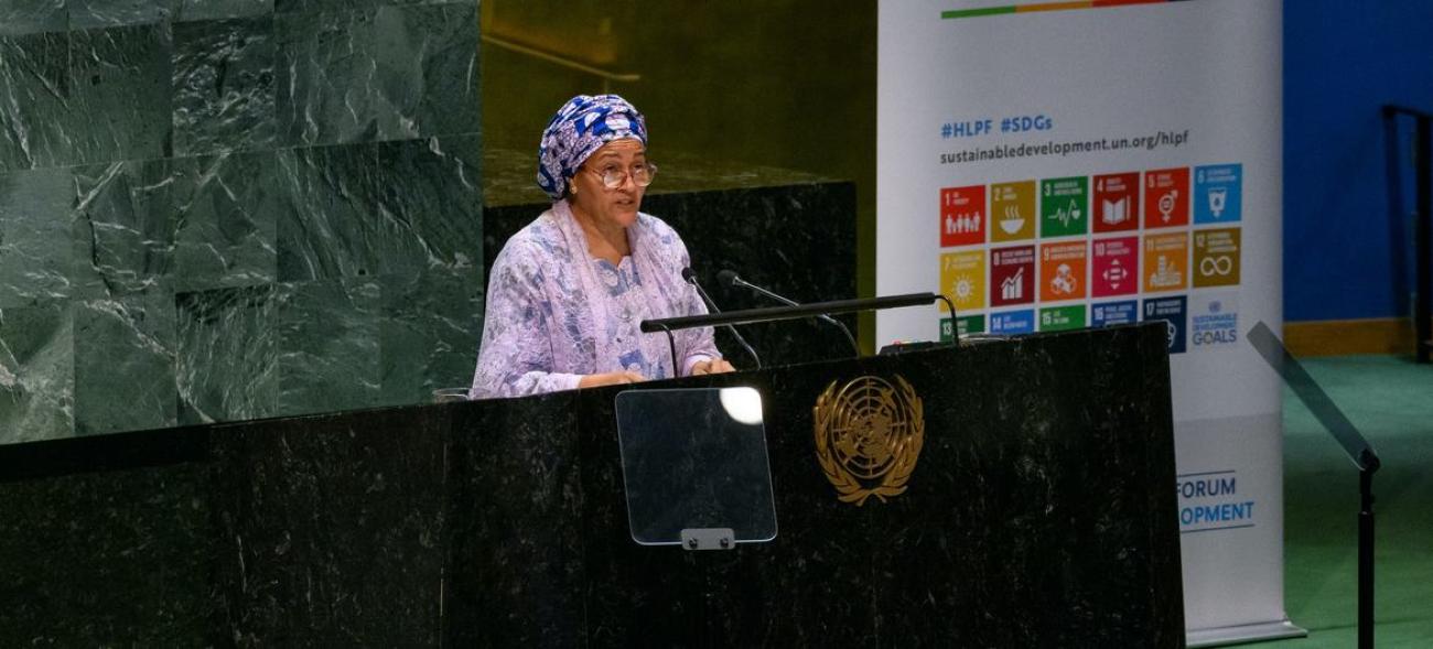 A woman in a lavender dress and headscarf speaks into a microphone at a podium in the UN rotunda.