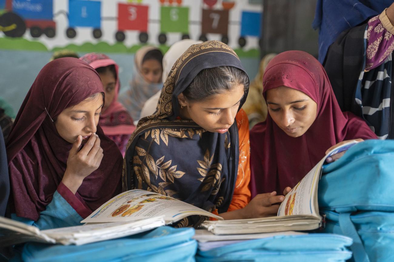 Three girls in red and brown dresses read over school textbooks in a classroom.