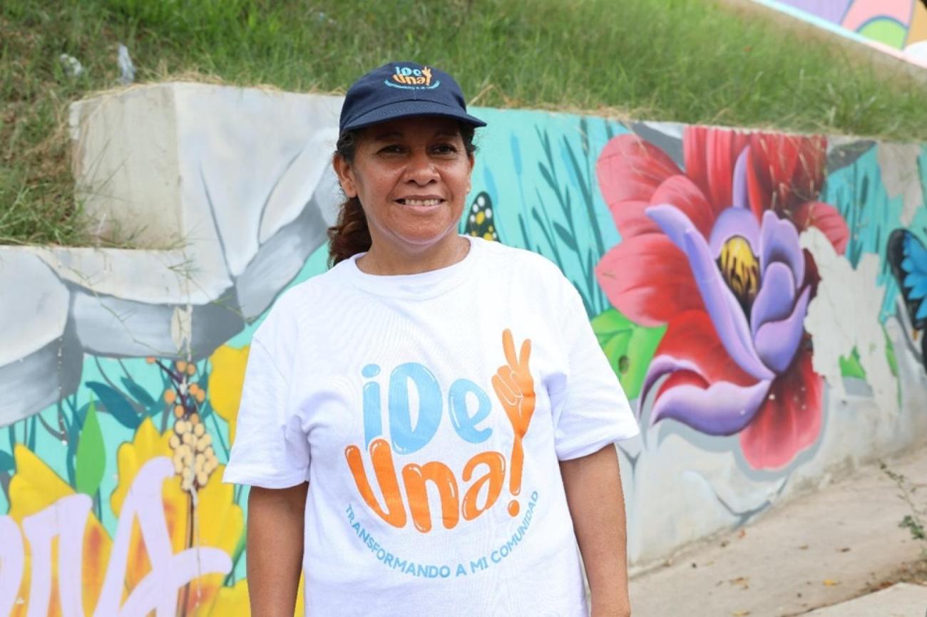 A woman in a colourful white shirt abd a cap stands in front of a painted wall.