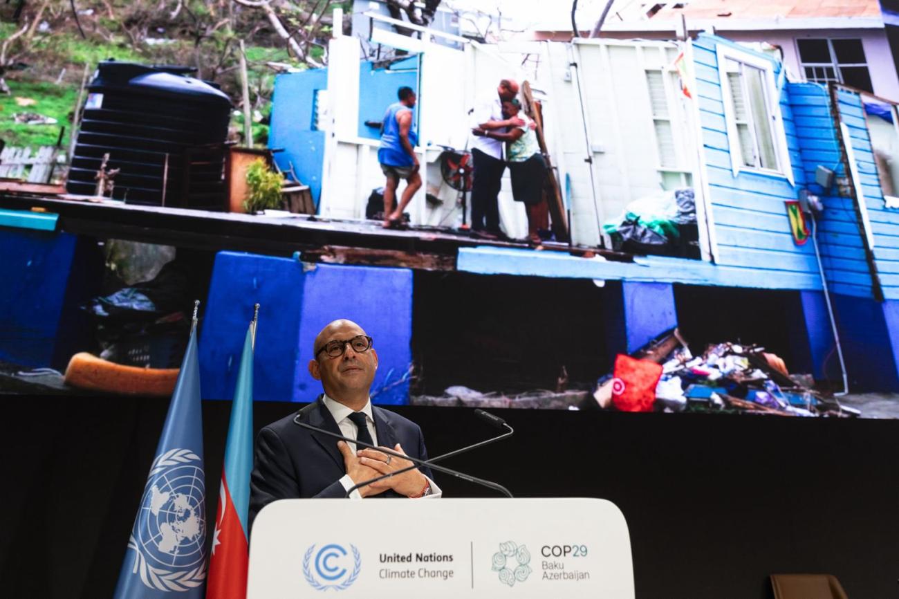 A man in a black suit addresses a crowd from a podium, while pictures of a disaster are projected on the screen behind him