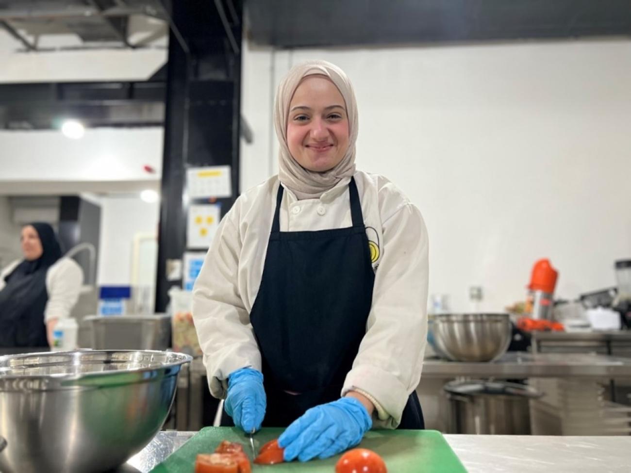 A woman in a white headscarf, black apron and blue gloves cuts vegetables on a kitchen counter.