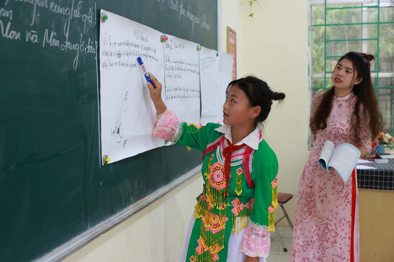 A girl in a colourful green dress points to a white board in a classroom while another woman in a pink dress, likely a teacher, watches on.