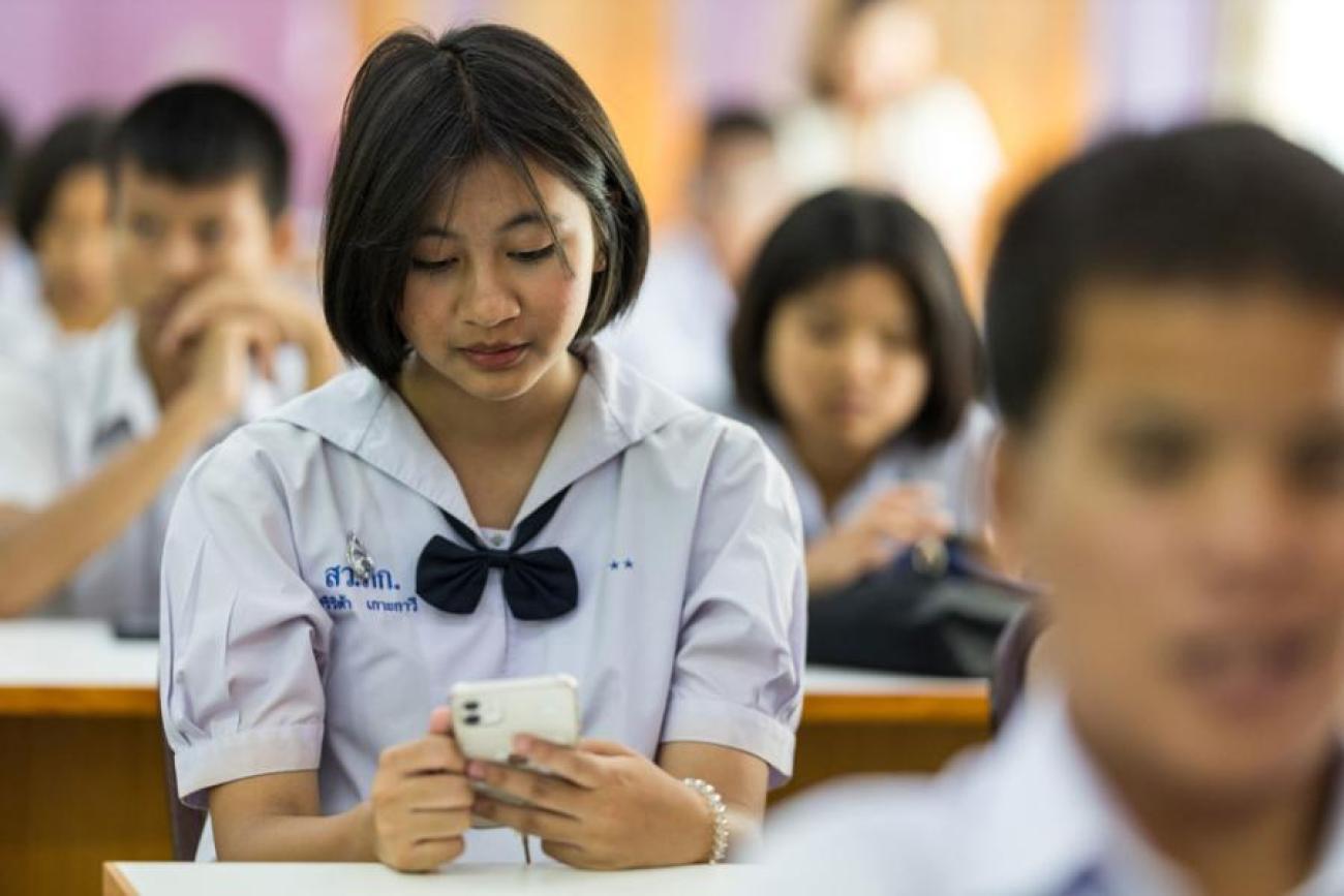 A girl student in a white school uniform checks her mobile phone while sitting in a classroom
