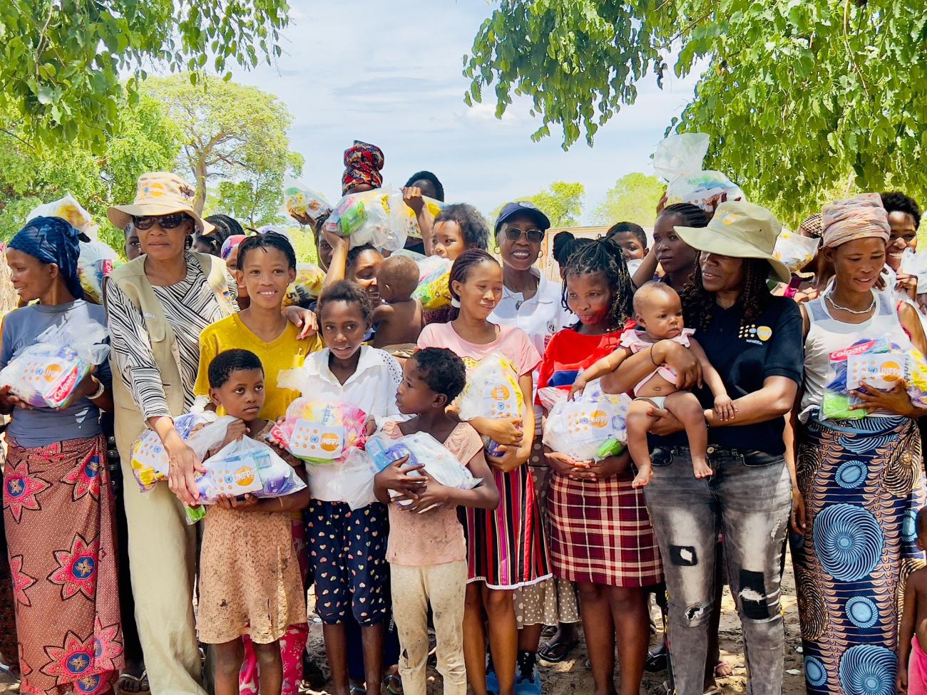 A group of women and children gathered, wearing colourful dresses and hats. In the midst of this group is a woman in a white shirt with glasses and a hat, the UN Resident Coordinator in Namibia