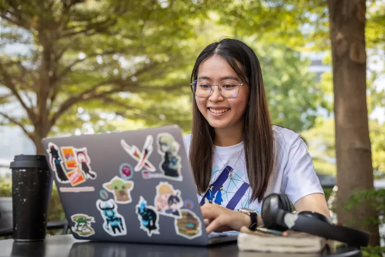 A girl in a white shirt wearing glasses smiles as she types into a laptop computer covered with stickers