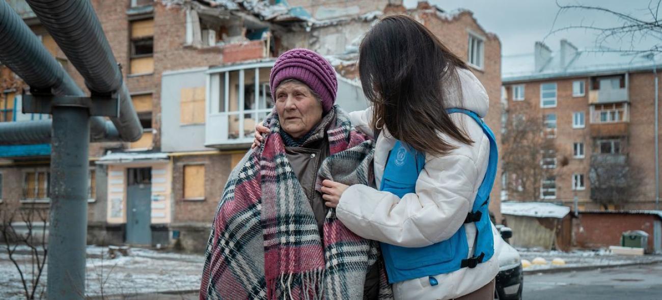 一位女性工作人员（右）搀扶着一位戴着紫色帽子裹着格子围巾的老年女性，站在被战乱破坏的房子前