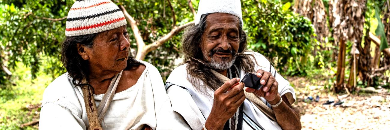 Two indigenous peoples, a man and a woman dressed in traditional clothes sitting side by side, the man in using a mobile phone