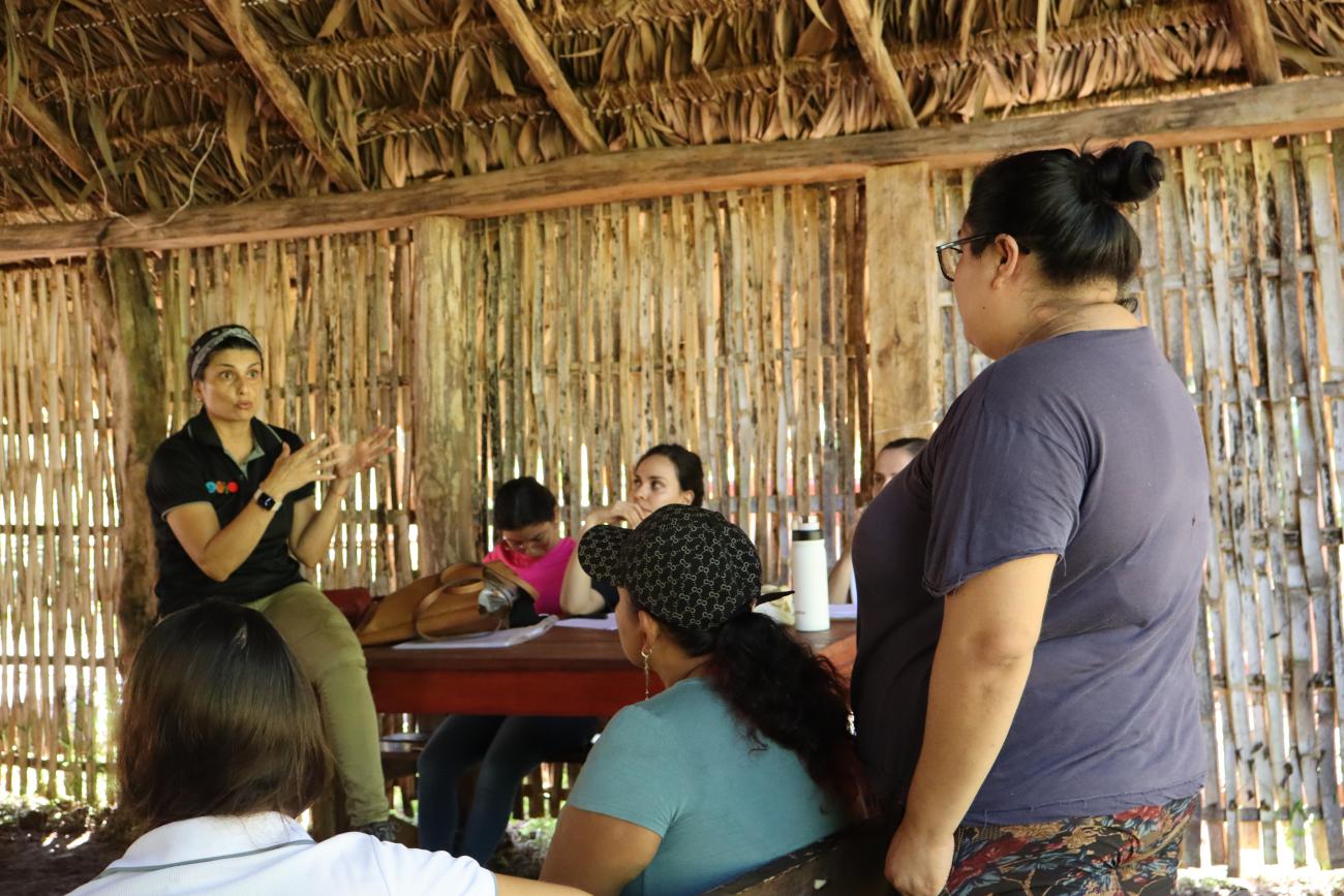 A woman in a black shirt speaks to two other women in a thatched house