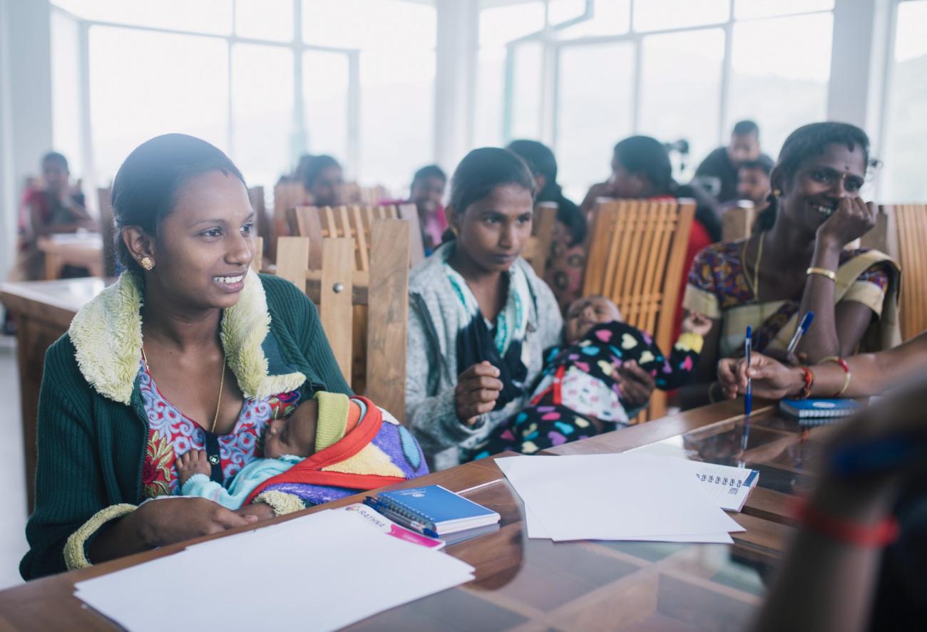 A row of women with children sit at desks while they fill out papers