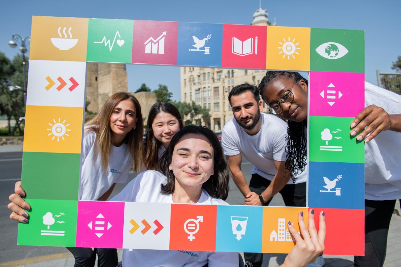 A group of volunteers, young men and women, hold up a square frame with the SDGs on it.