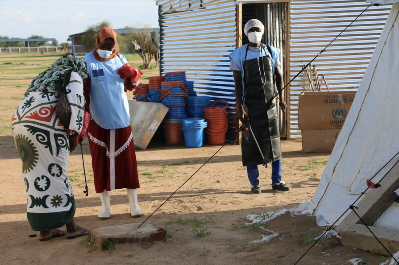 两名非洲妇女和一名非洲男性站在黄土地上，身旁是简易的房屋。